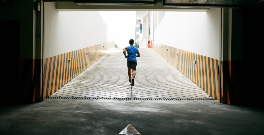 Black-haired athlete listening to music while running out of underground parking lot, back view